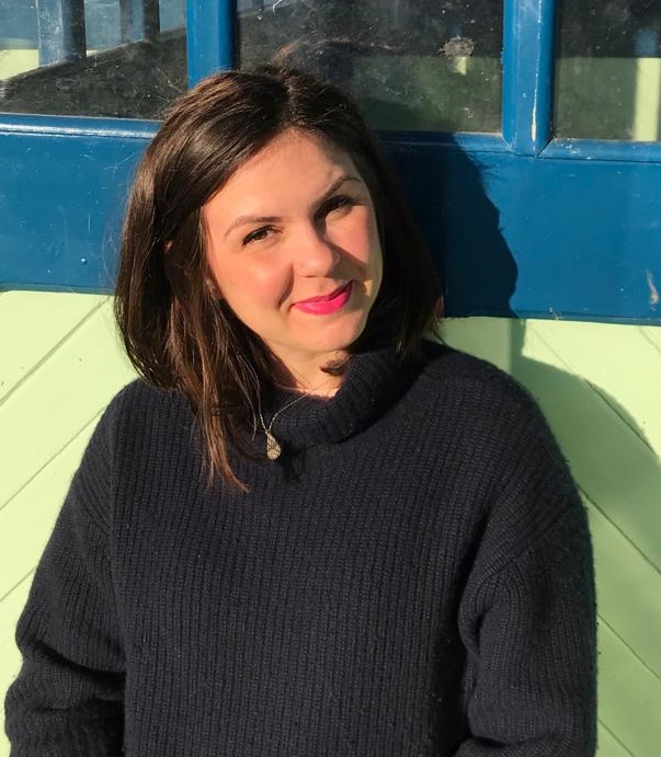 Headshot of young, white smiling woman. She wears a black jumper, lipstick and has mid-length brown hair.