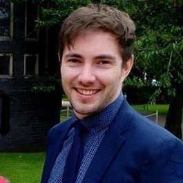 Headshot of smiling, young, white man in navy blue suit. He has brown hair and facial hair and is outside.