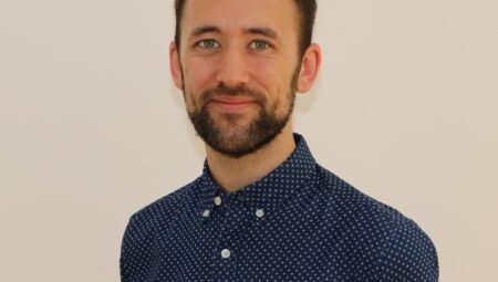 Headshot of Oliver - young white man with brown hair, brown beard and mustache wearing a navy blue shirt.