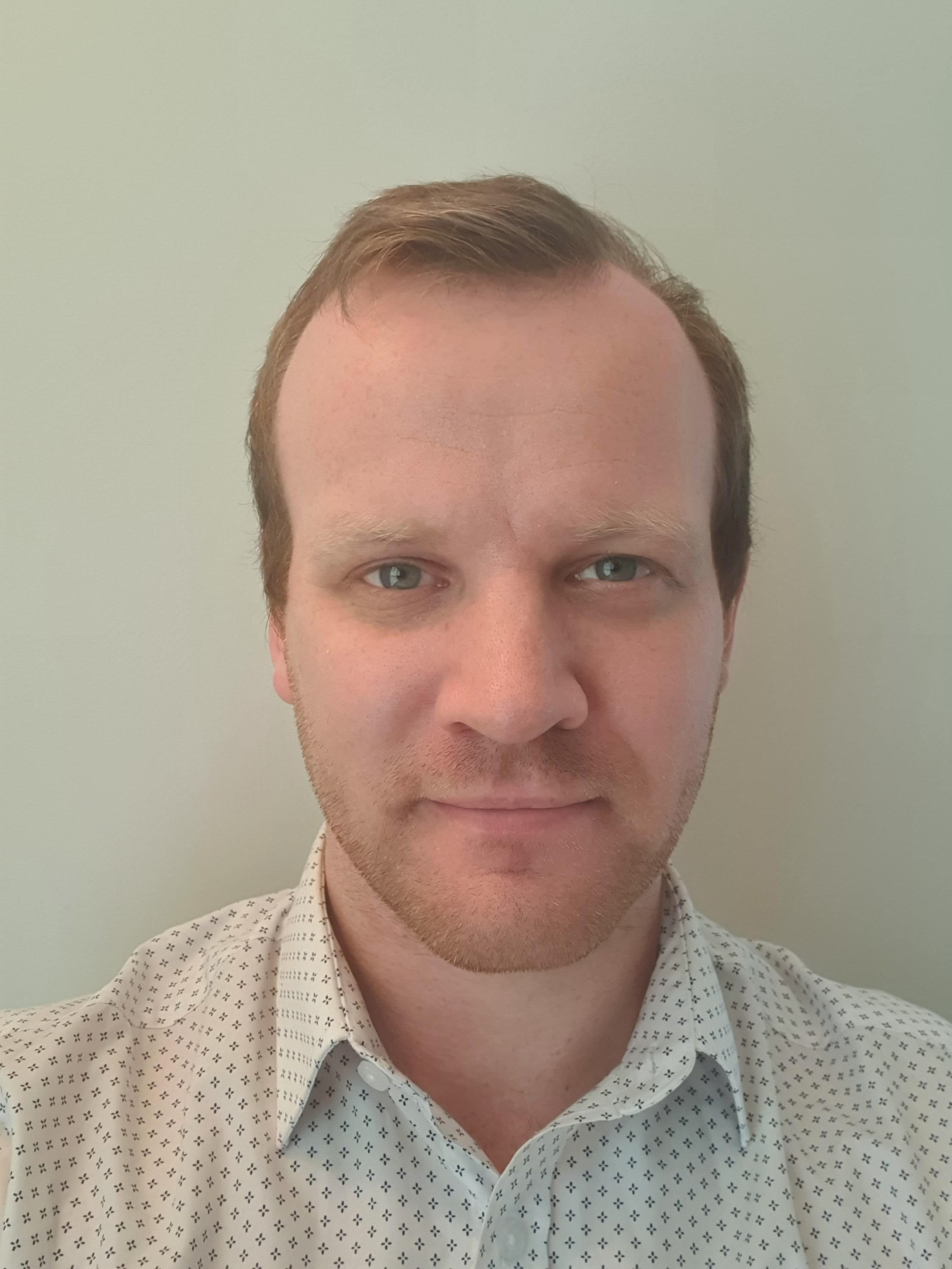 Headshot of man with ginger hair and facial hair, wearing white shirt.