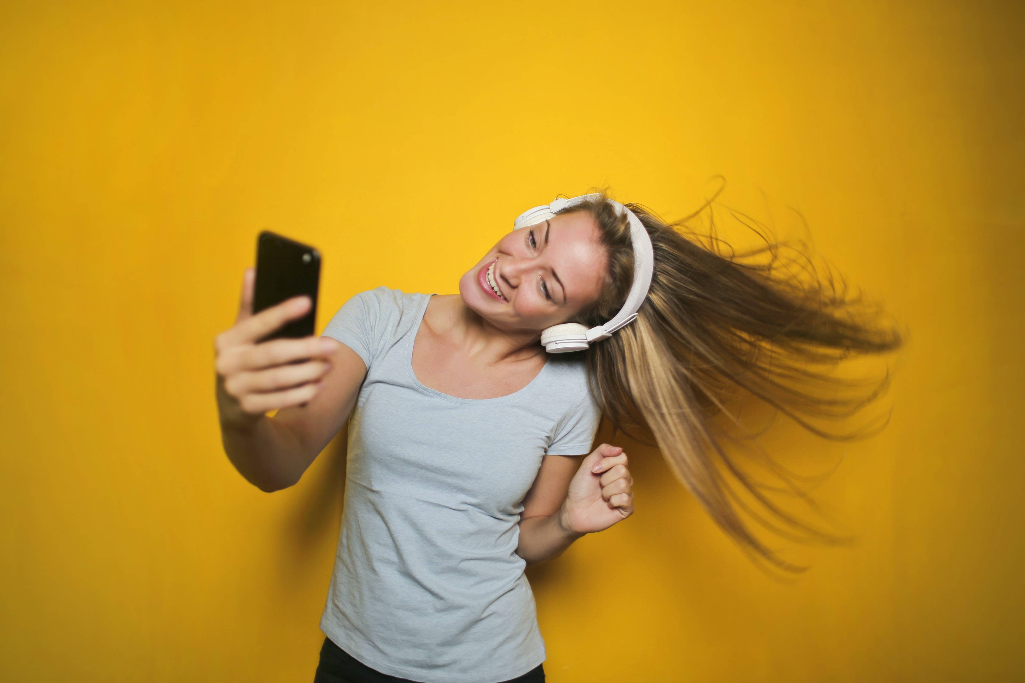 Young woman with headphones on is looking at her mobile phone and dancing, flicking her hair to one side. Mustard yellow backdrop.