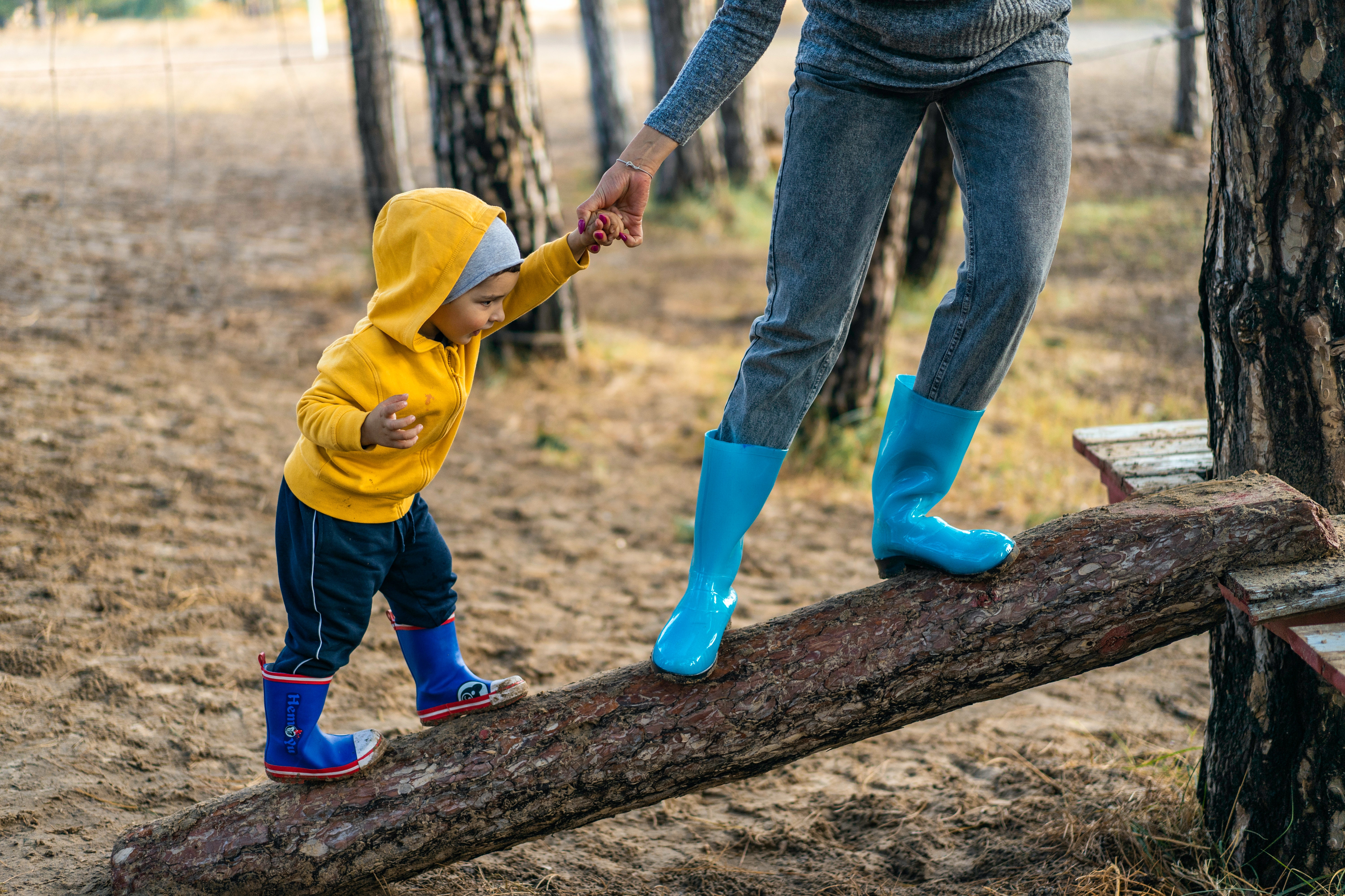 Child with blue wellies and bright yellow hooded top is stood, balancing, on a tree trunk. The child holds an adults hand who we can see only from the waist down. The adult wears light blue wellies. 